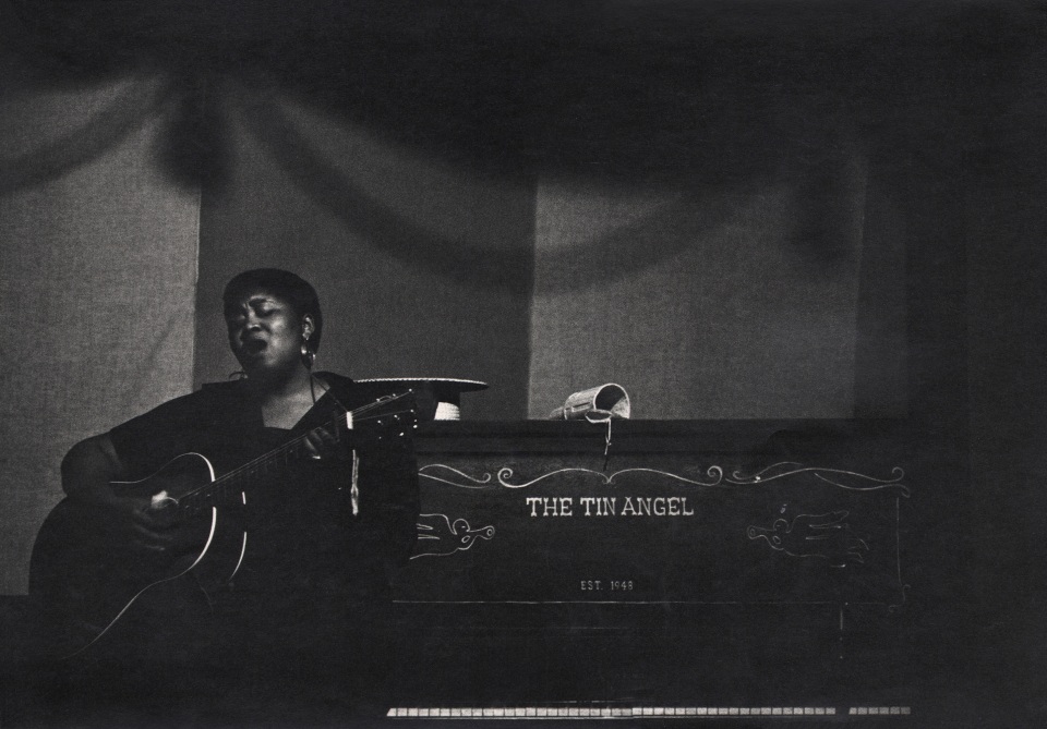 Bob Willoughby, Folksinger Odetta, Tin Angel Nightclub, San Francisco, Calif., USA, ​1954. Subject sits holding a guitar and singing on stage in front of a piano marked &quot;The Tin Angel&quot;