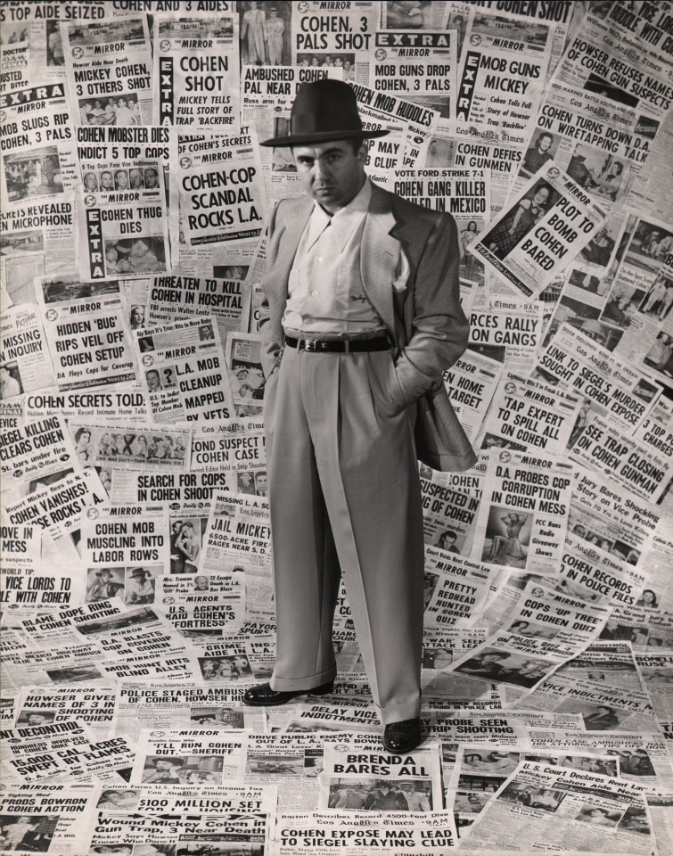 24. Ed Clark, Mickey Cohen, Gangster, 1949. A suited man stands against a backdrop of newspapers pasted to the wall and ceiling, hands in pockets, looking to the camera with a serious expression.