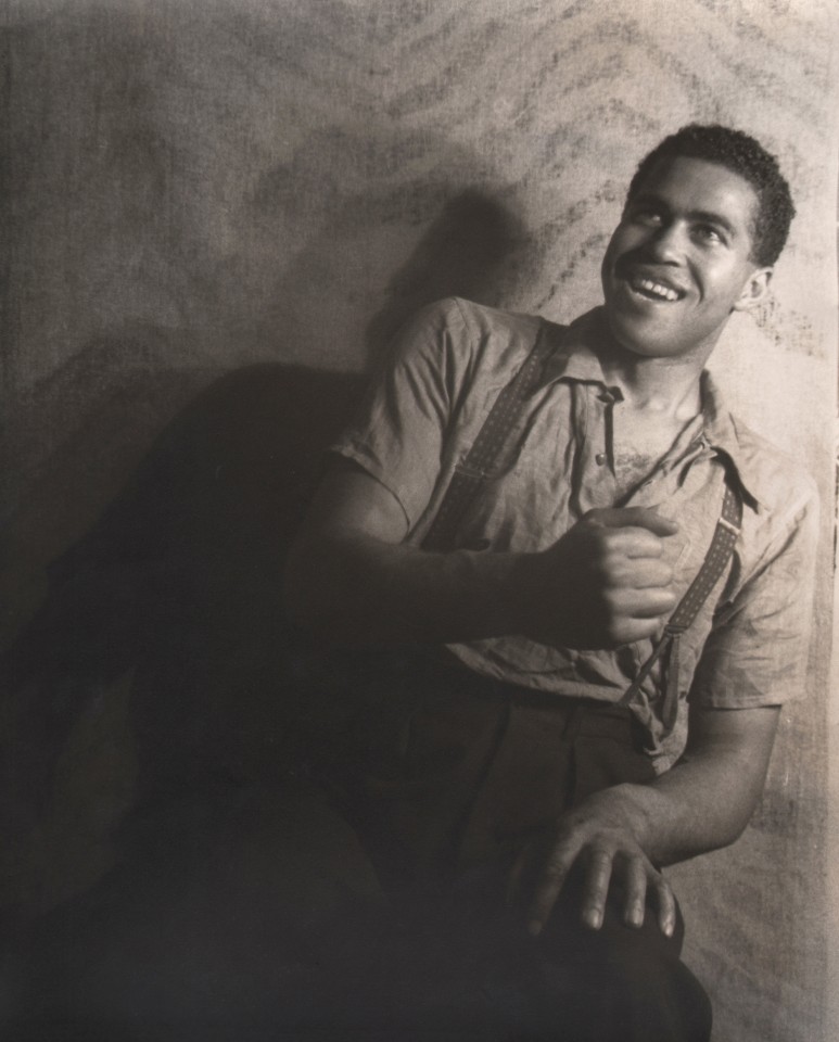 17. Carl Van Vechten, Robert Earl Jones in Langston Hughes' play, 1938. Seated portrait with subject smiling up and to the left of the frame, one hand in a fist in front of him.