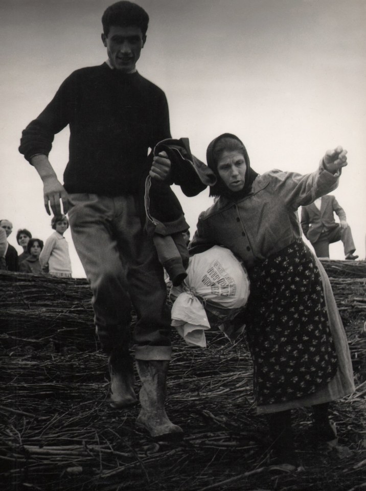 Pietro Donzelli, Flood in Polesine, ​1960. Two figures walk downhill towards the camera. Other figures watch from the background at the top of the hill.
