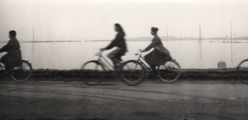 Pietro Donzelli, Flood in Polesine, ​1960. Three figures on a seaside road ride bicycles from right to left across the horizontal frame.