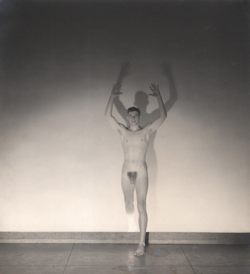 George Platt Lynes, Jeremy Jackson, ​1935. Male nude figure stands with arms and one leg raised against a spot-lit wall.