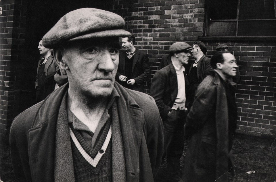 27. Patrick Ward, One of a series of pictures to do with a slump in shipbuilding along the Clyde, ​c. 1966. A group of men standing outside of a brick building. One figure in a hat and jacket is in the foreground left.