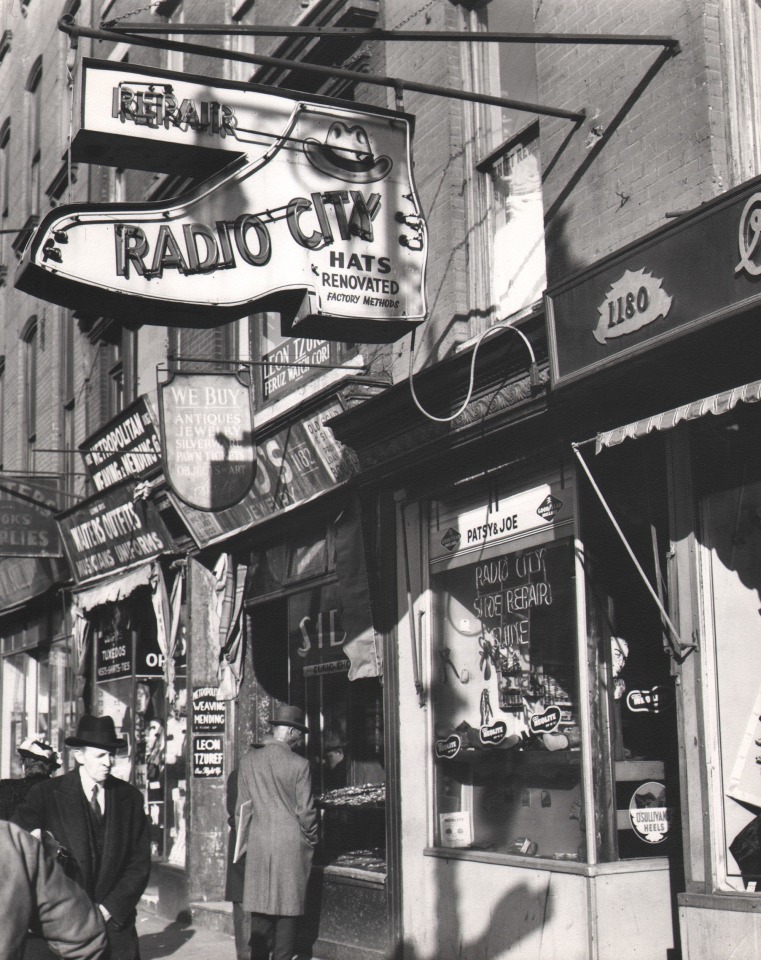 3. Todd Webb, Rockefeller Center, New York City. Shops on Sixth Avenue near Rockefeller Center, ​1947. Street scene featuring a shoe-shaped neon sign for &quot;Radio City Hats&quot; in the upper left of the frame.