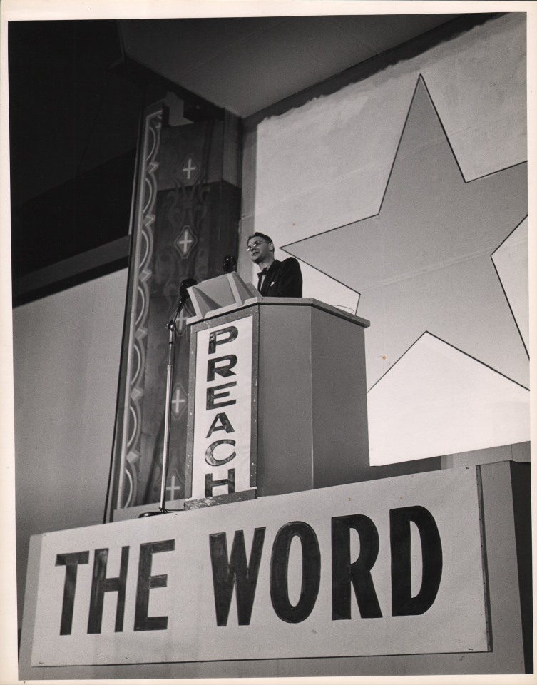 32. Gordon Coster, Preach the Word, ​c. 1940. A suited man stands at a raised podium that reads &quot;Preach The Word&quot;. A large star is painted on the wall behind him.