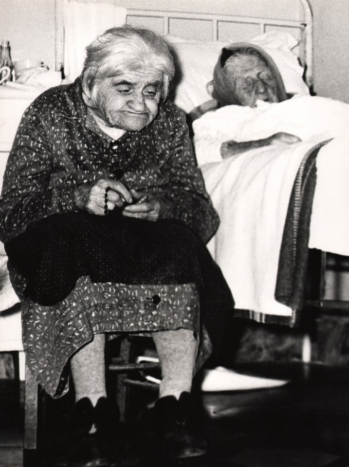 03. Mario Giacomelli, Verr&agrave; la morte e avr&agrave; i tuoi occhi, 1966&ndash;1968. High contrast image. One woman sits with a blanket and rosary in her lap in the foreground, another sleeps in bed in the background.