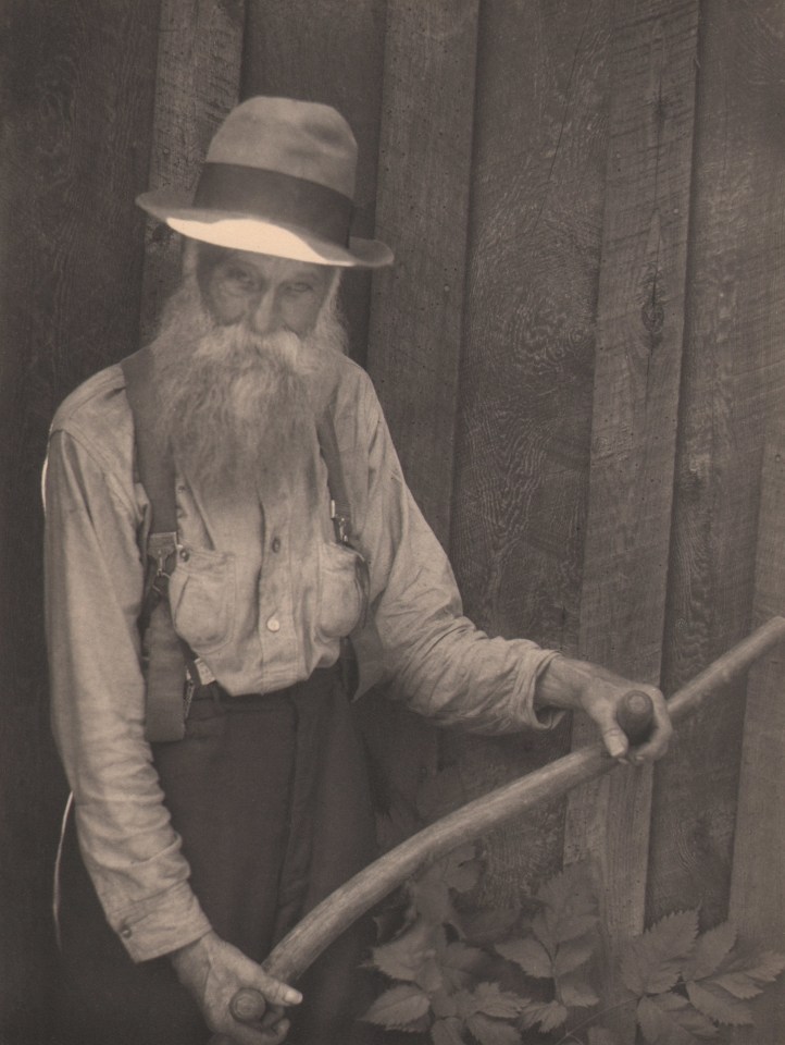 Doris Ulmann, Untitled (Farmer holding oxen yoke), ​1928&ndash;1934. Older bearded man in a hat standing against a wooden wall and suspenders holding a wooden rod with two handles.