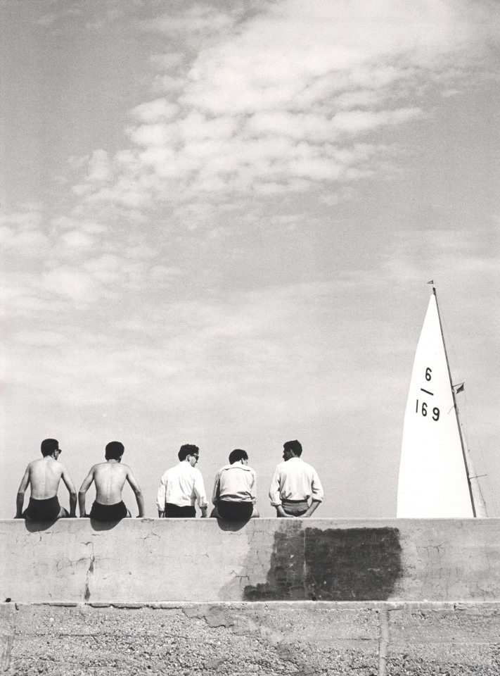 Nino Migliori, Quattro Con, 1955. Five men seated on a wall photographed from the back. A ship's sail is in the right of the frame.