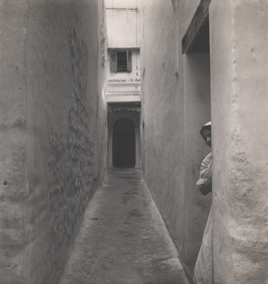 51. Cecil Beaton, Tangier, c. 1945. View down a narrow alleyway; a figure peeks out of a doorway on the right.