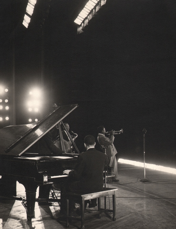 Bob Willoughby, Miles Davis, Shrine Auditorium, Los Angeles, CA, ​1950. Subject stands on stage playing the trumpet in front of a piano and other accompanying musicians.