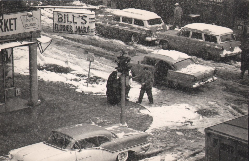 44. W. Eugene Smith, As From My Window I Sometimes Glance, ​1957&ndash;1958. Elevated view of a snowy street scene. A man helps a group of women from the sidewalk corner to a car. Sign on the corner of the nearest building reads &quot;Bill's Flower Mart&quot;