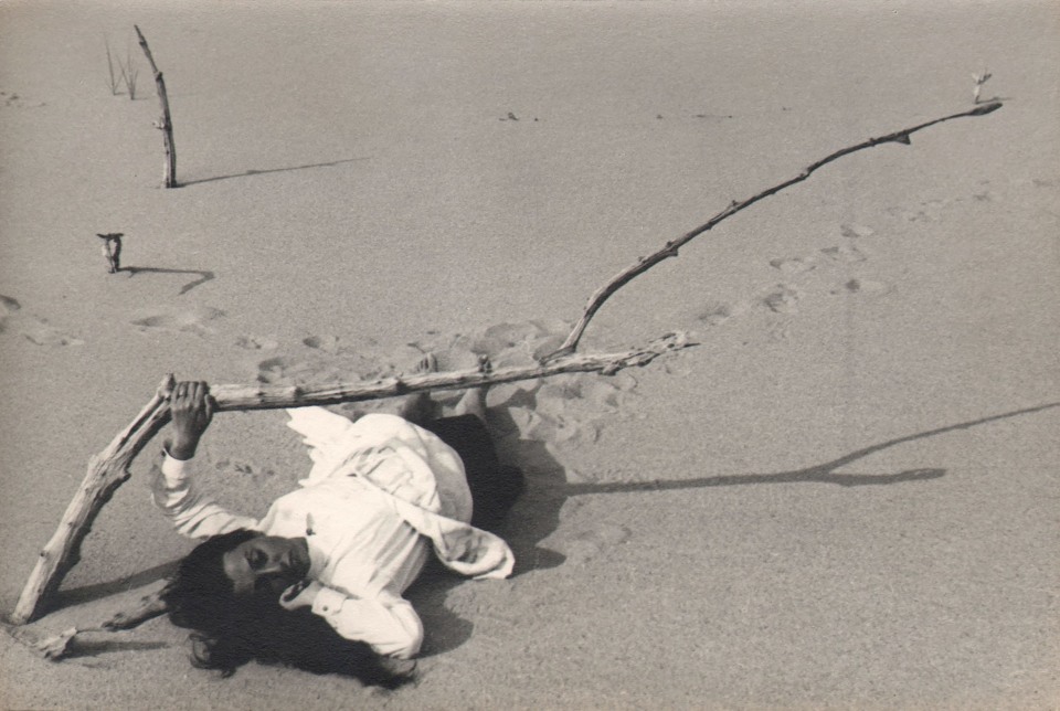 PaJaMa, Fidelma Cadmus, ​c. 1945. A woman in white lays in the sand beneath a dried wooden branch extending from the lower left to the upper right of the frame.