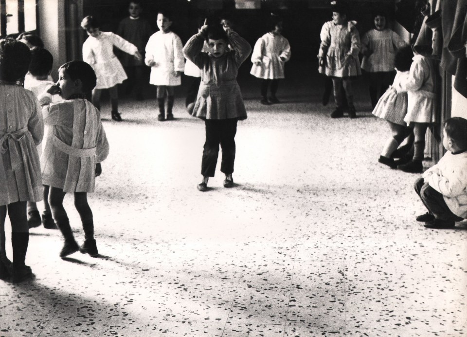 23. Renzo Tortelli, Piccolo Mondo, 1958&ndash;1959. A young boy stands in the upper center of the frame with fingers pointed above his head like horns. Children line the walls around him.