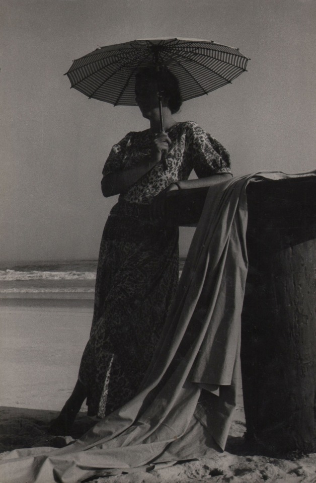 PaJaMa, Margaret French, ​c. 1945. A woman stands on the beach holding a parasol, leaning against a wooden structure with fabric draping from it.