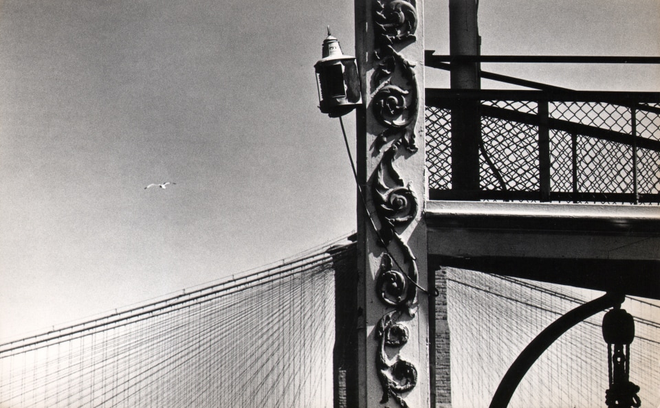 8B. Jan Lukas, New York City, Brooklyn Bridge, ​1964. Detail of an ornate metal beam in the foreground that aligns with the vertical towers of the Brooklyn Bridge in the background.