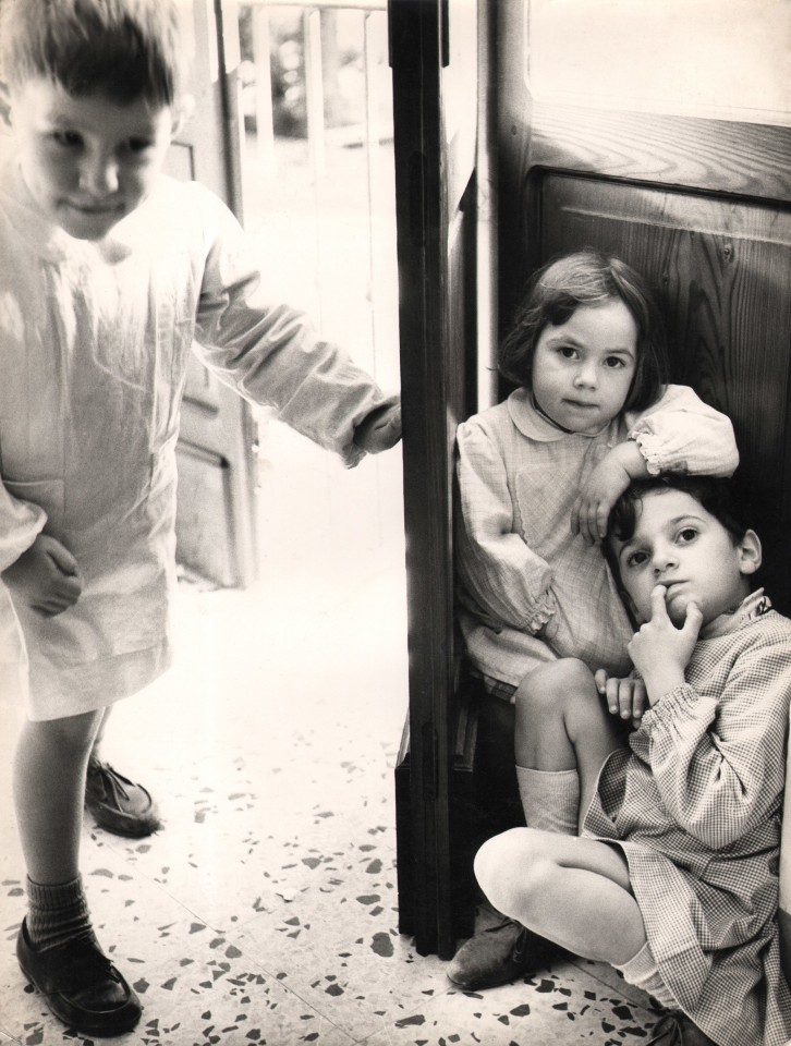 15. Renzo Tortelli, Piccolo Mondo, 1958&ndash;1959. High contrast image. A boy walks through a doorway on the left of the frame; two children seated on the floor on the right.