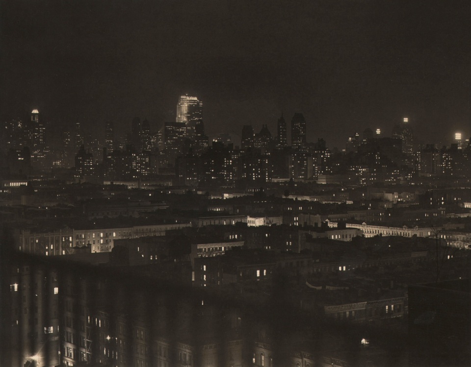 Paul J. Woolf, Rockefeller Center Looking Southwest, ​c. 1936. Night time city view from a balcony or rooftop with an out-of-focus railing in the lower left of the frame.