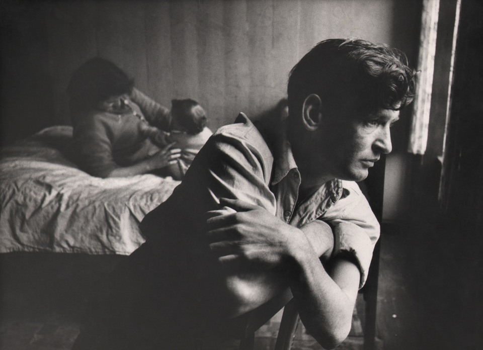 37. Bert Hardy, Untitled, c. 1957&ndash;1960. A seated man in the foreground looks toward sunlight coming from the right of the frame. In the background left, a woman and baby play on a bed.