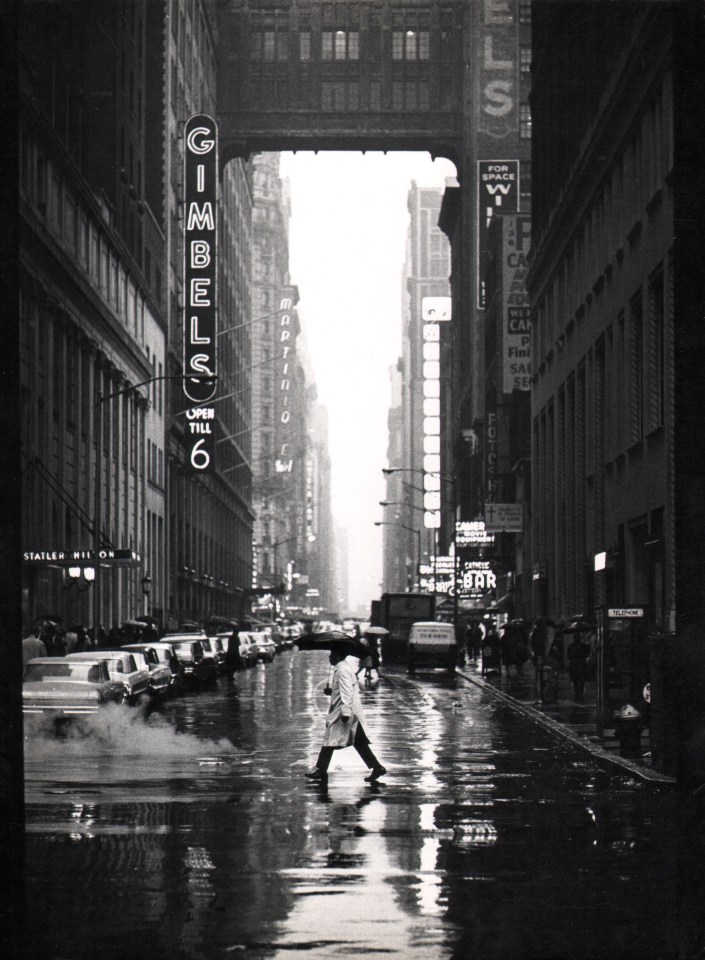 10. Jan Lukas, New York, 32nd Street, ​1964. A man with a trenchcoat and umbrella walks across a wet car-lined street. The neon sign for Gimbels is on the left midground.