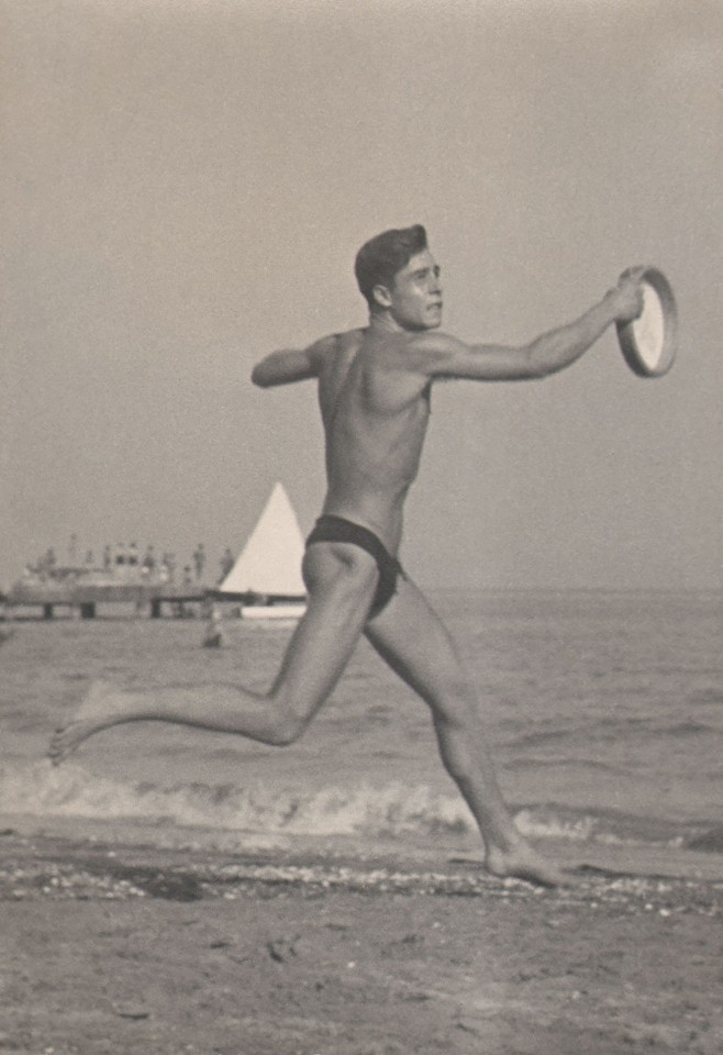 PaJaMa, Silvano Gasparini, Venice, ​c. 1949. A man in a swimsuit catches a frisbee, running from left to right across the frame. The ocean and a sailboat are in the background.