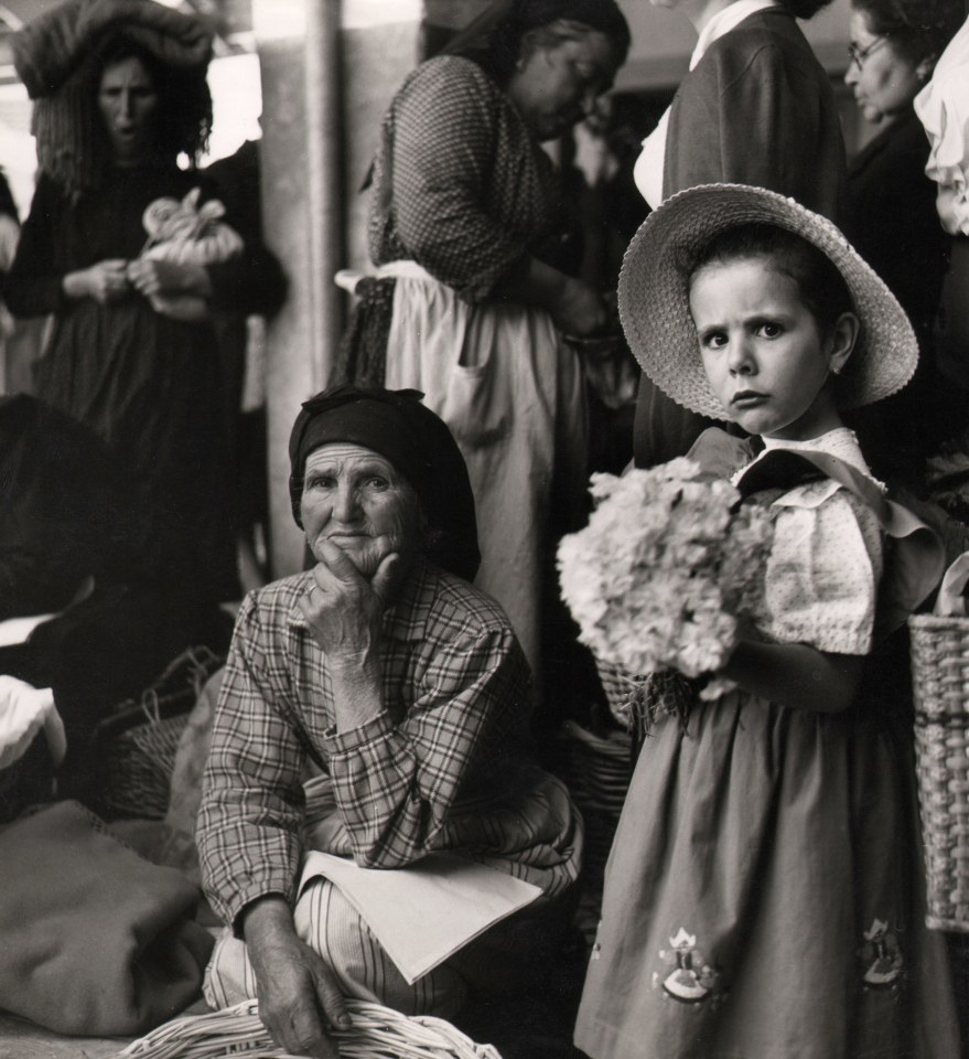 42. Sabine Weiss, Young and old faces from the north of Portugal, c. 1950. A young girl holding flowers stands in the right of the frame, looking to the camera with a concerned expression. An older woman is seated on the left of the frame, also looking to the camera with one hand on her chin.