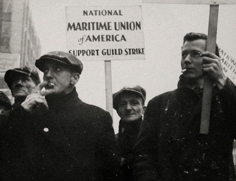 Gordon Coster, W.P.A. Parade, Chicago, 1939. Four men walk in black coats on a snowy day, smoking cigarettes. One holds a sign that reads &quot;National Maritime Union of America Support Guild Strike&quot;