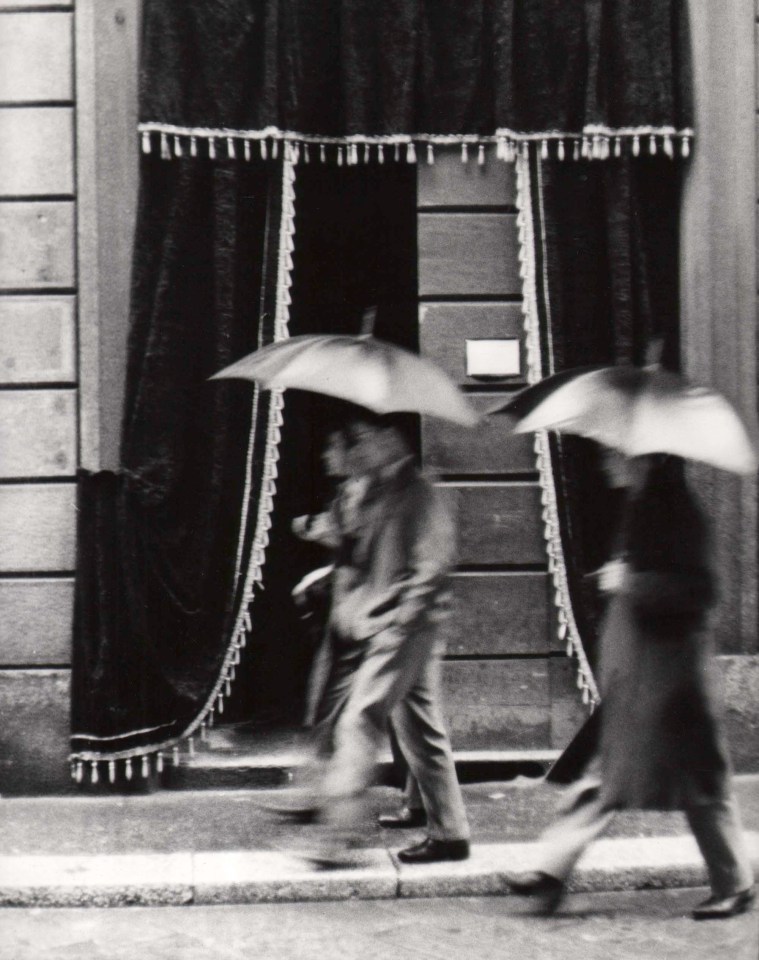 Mario Carrieri, Milano, ​c. 1958. Three figures, blurred with motion, walk across the frame right to left, holding umbrellas. Black tasseled curtains hang from the building behind them.