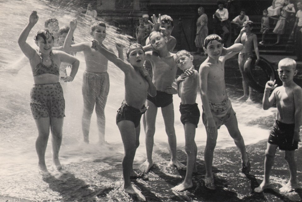 13. Jeanne Ebstel, Untitled, ​c. 1947. Group of children in swimsuits on the street, being sprayed with water gushing from out of the frame.