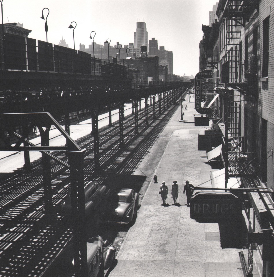 33. Esther Bubley, View of Third Avenue El looking downtown from 53rd Street. The El goes as far downtown as the Battery, 1946. Elevated view of train tracks and three pedestrians walking on the sidewalk on the right of the frame.