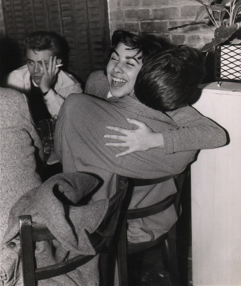 23. Weegee, James Dean in Greenwich Village, ​c. 1955. Two seated figures in the foreground embrace while Dean holds his hand to his face with a cigarette in the background left.