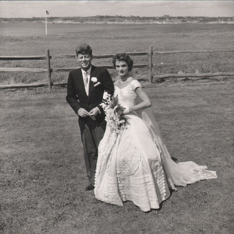 47. Toni Frissell, John F. Kennedy &amp; Jacqueline Bouvier on Their Wedding Day, Newport, Rhode Island, 1953. The couple pose with arms linked in the middle of the frame, a wooden fence and vast field in the background.