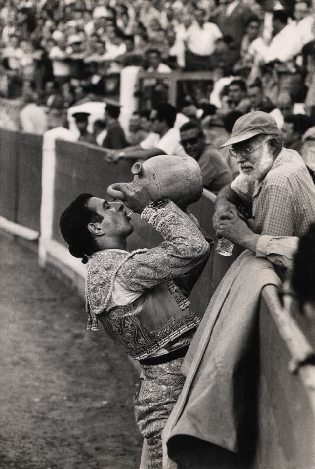 42. Larry Burrows, Bullfighting (Hemingway), 1959. A bullfighter at the perimeter of the fighting ring drinks from a ceramic vessel as Hemingway looks on from the other side of the wall.