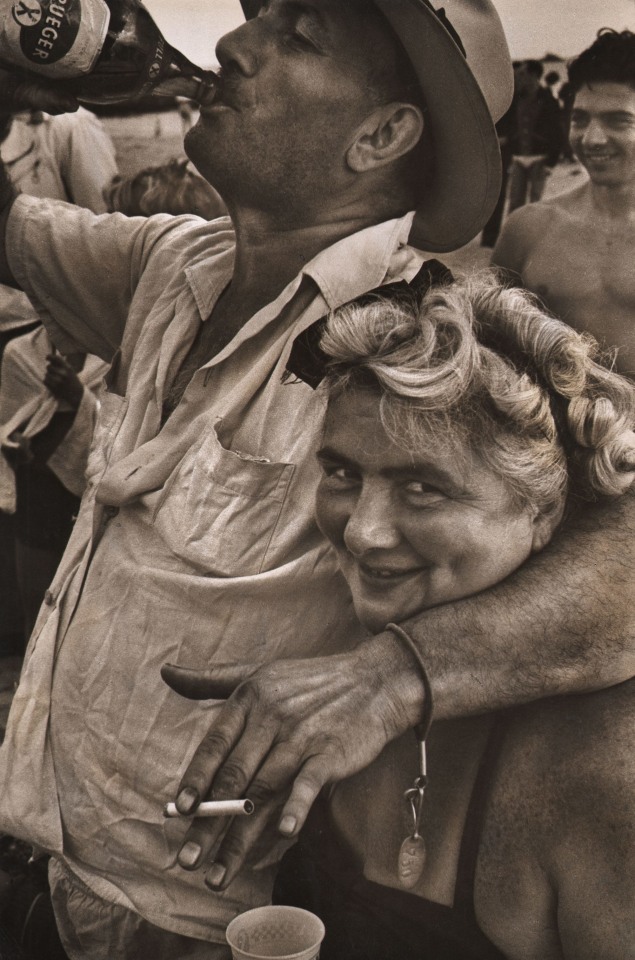 Harold Feinstein, Man and wife drinking Krueger beer, Coney Island, ​1952. A man holds a cigarette in one hand and drinks beer with the other, with one arm around a woman who smiles at the camera.