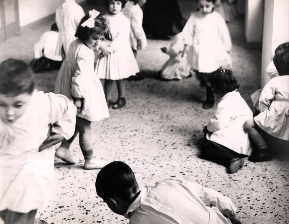 24. Renzo Tortelli, Piccolo Mondo, 1958&ndash;1959. High contrast photo. A haphazard circle of children in white, sitting or standing on a speckled floor.