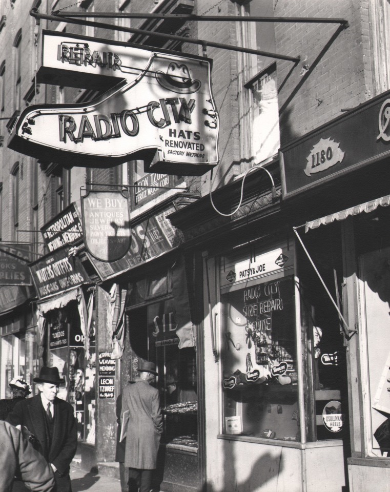 56. Todd Webb, Shops at Sixth Avenue near Rockefeller Center, 1947. Street-level view of pedestrians outside a shop with a shoe-shaped sign that reads &quot;Radio City Repair - Hats Renovated Factory Methods&quot;