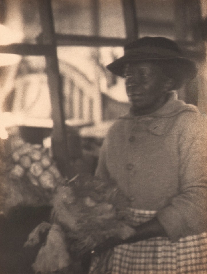 Doris Ulmann, Untitled (Vegetable seller), ​1928&ndash;1934. Woman in a hat and checkered skirt stands holding vegetables and looking to the left of the frame.