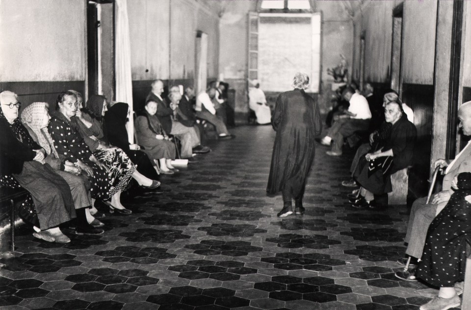 05. Mario Giacomelli, Verr&agrave; la morte e avr&agrave; i tuoi occhi, 1966&ndash;1968. High contrast image. A woman walks down a hallway, back to the photographer. The hall is lined with seated women.