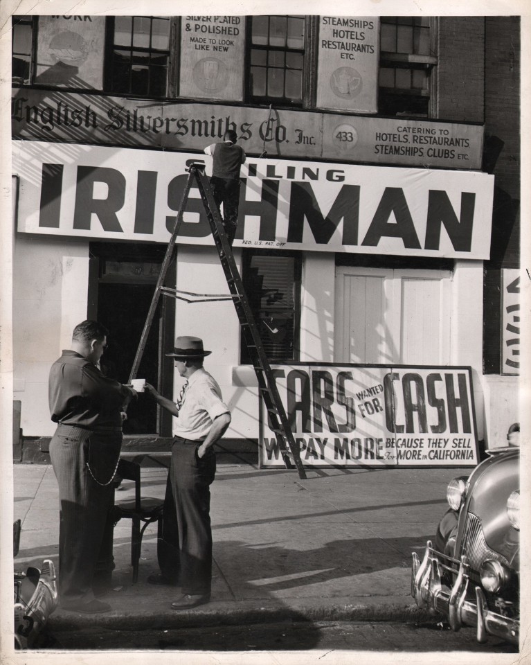 13. Weegee, Untitled, ​c. 1945. Two men stand in the foreground left talking; a third man stands on a ladder in the background in front of a large sign that reads &quot;Irishman&quot;