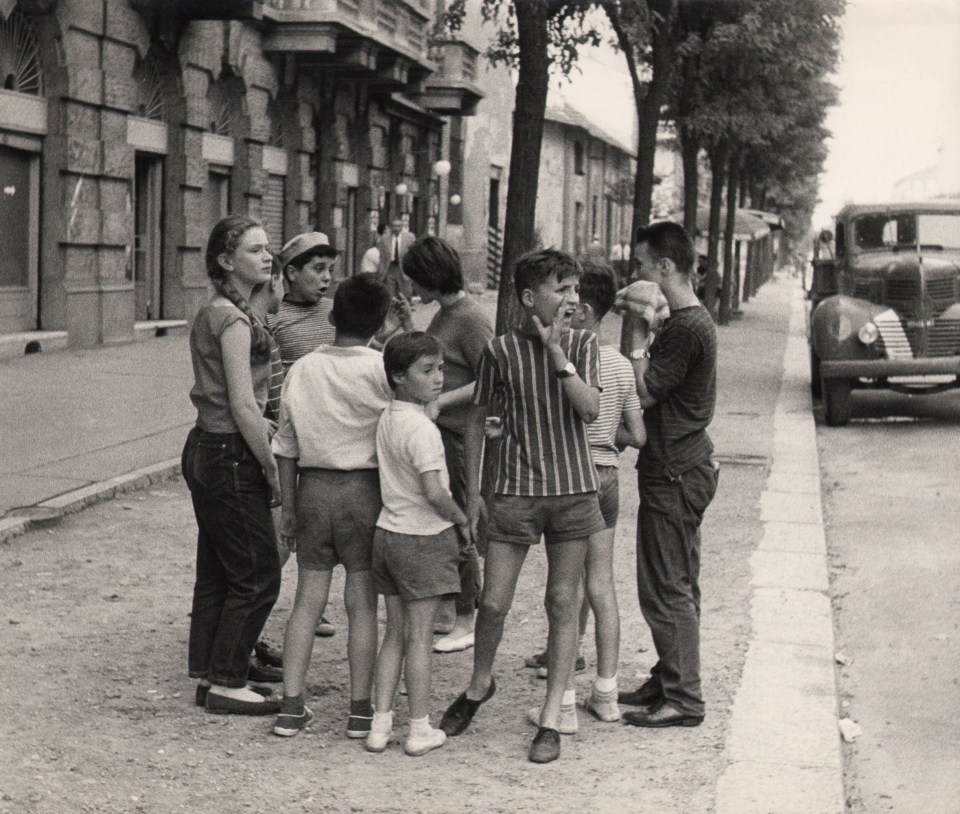 Mario Finocchiaro, Ragazzi di Milano, ​1959. A group of children gather on a tree-lined street. Most are looking off the the right of the frame and one boy is yelling in that direction.
