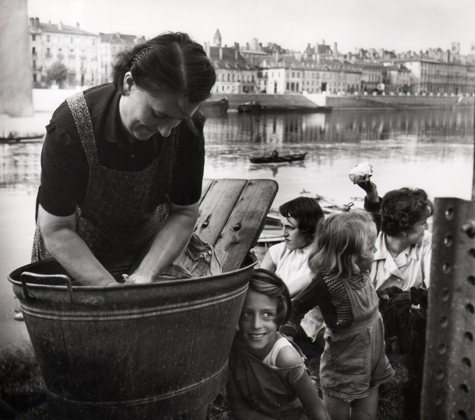 50. Janine Ni&eacute;pce, Untitled, c. 1960. A woman in the foreground left washes clothing in a tin bucket; children play behind her to the right. A river, canoe, and large buildings are in the background.