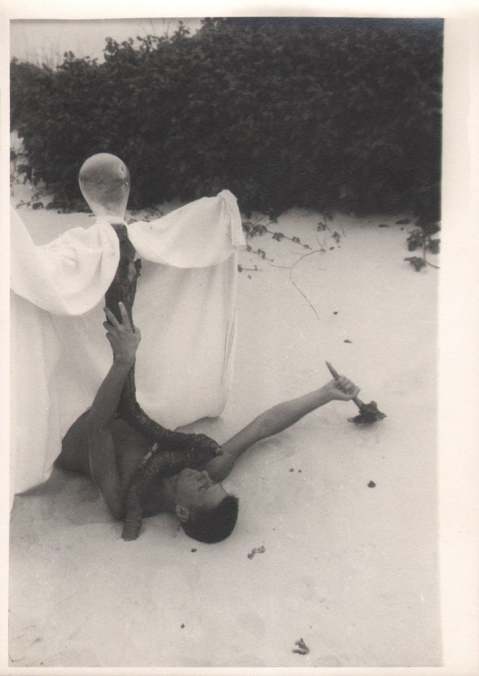 PaJaMa, Paul Cadmus, c. 1945. Subject lays in the sand holding a vaguely figural sculpture above him made of driftwood, a white sheet, and what looks like a balloon.