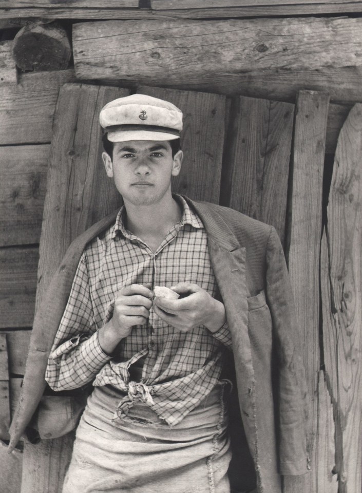 Tranquillo Casiraghi, Cavatore d'ardesia, ​1959. A young quarryman in a white cap, jacket draped over his shoulders, stands in front of a wooden structure.