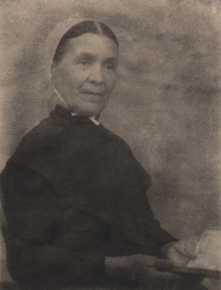 Doris Ulmann, Untitled (Shaker lady), ​1928&ndash;1934. Seated woman in a bonnet and black clothing holds open a small book in her lap, looking slightly to the right of the frame.