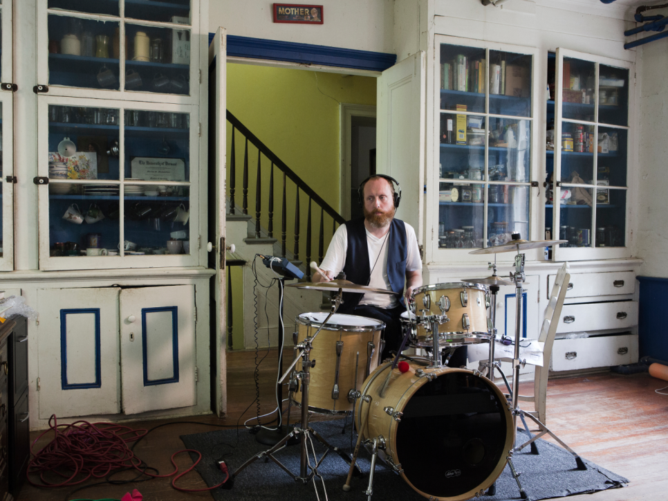 A bearded man playing drums in front of white cupboards with blue trim