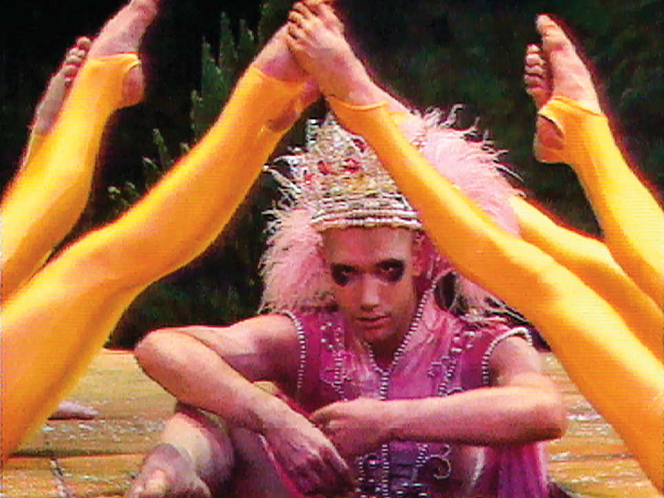Video still of a dance performance: man in pink costume under 7 legs with yellow tights