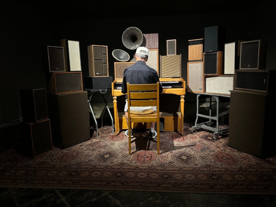 Person sitting at an organ in front of over a dozen mixed speakers 