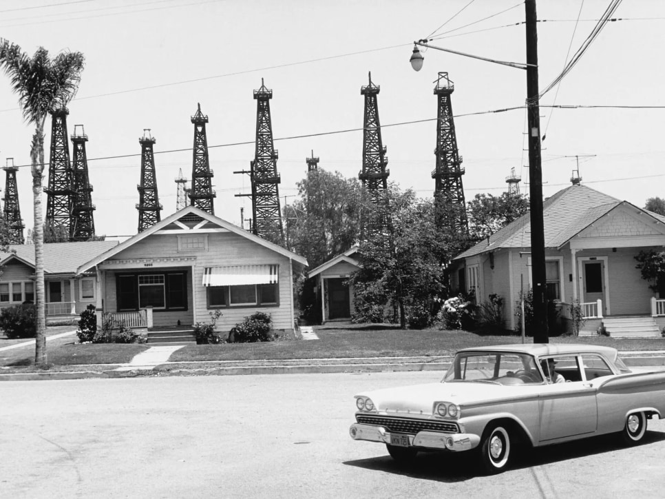 Black and white photo of a street, including a car, 2 houses and palm trees