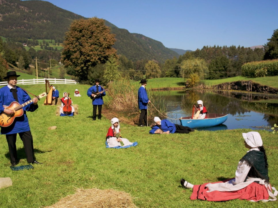 10 figures in folk clothing and musical instruments, standing in a grassy field by a small lake