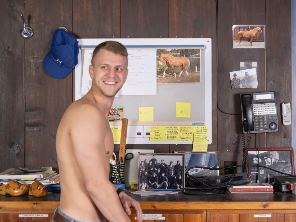 Shirtless young white man in front of an office wall with phone and photographs
