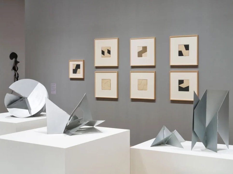 A Lygia Clark installation: 4 metal sculptures on plinths with 7 black and white collages hung on a grey wall in the background